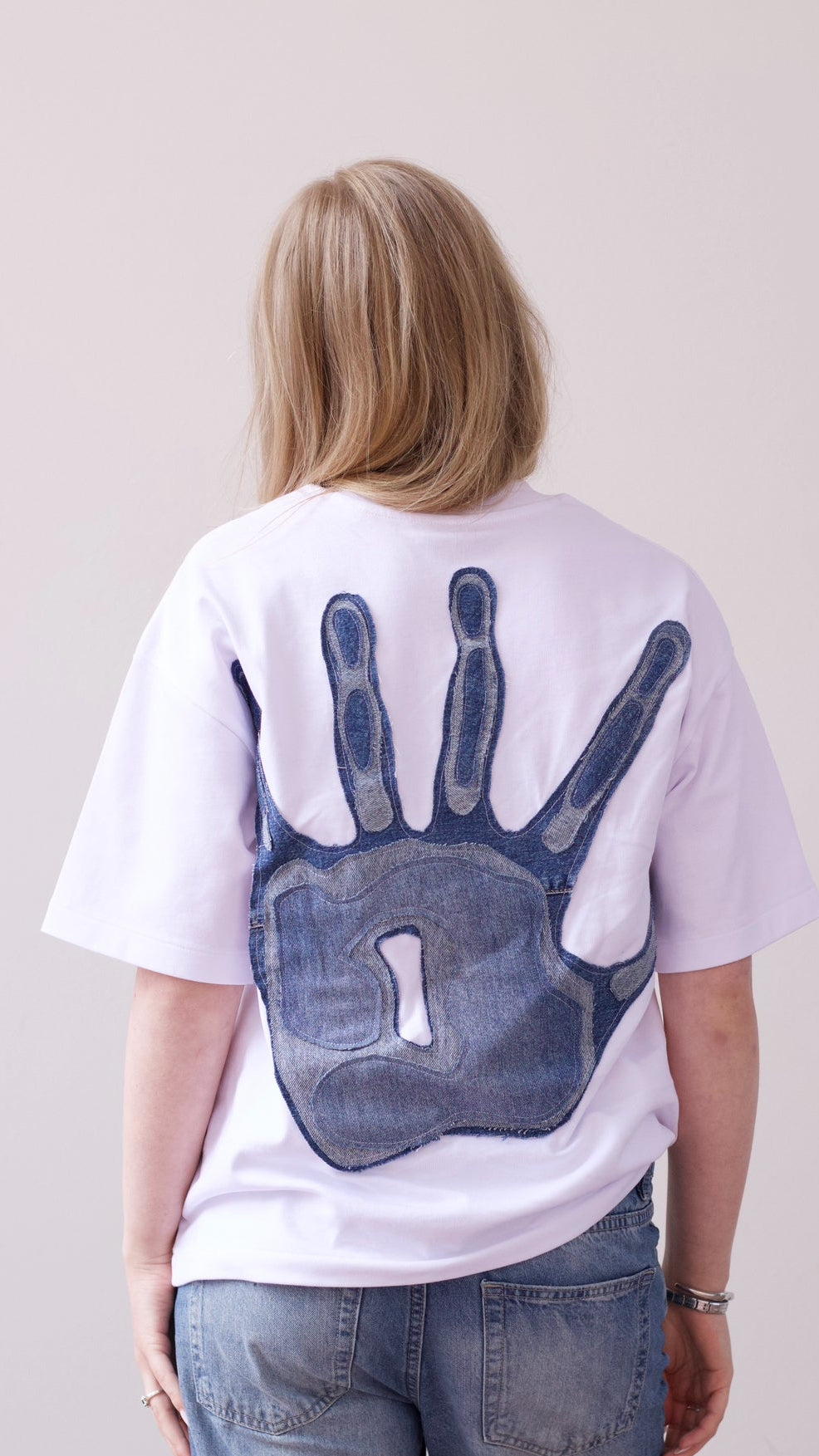 A woman viewed from behind wearing a white t-shirt with a large blue hand-shaped graphic on the back, paired with blue jeans, against a plain light beige background.